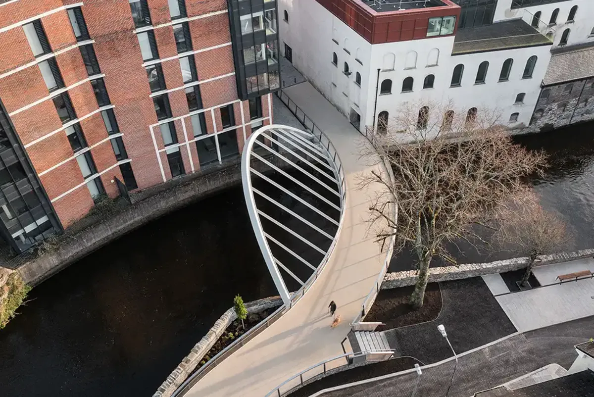 Aerial view of one of the Beamish and Crawford Bridges over the River Lee, showing a person and dog walking across the bridge, with buildings and a tree on the quays.