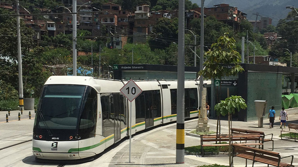 A train on street level tracks with park benches and houses on a hill in the distance