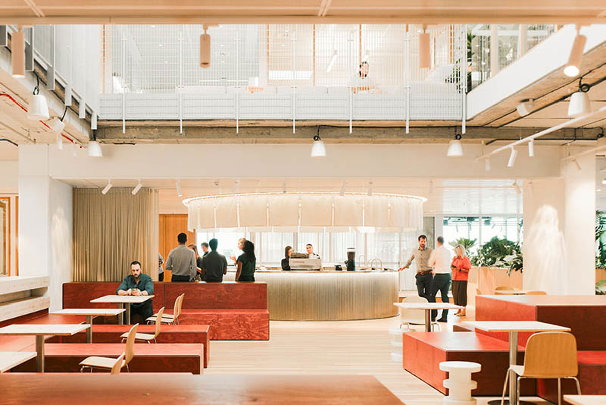 Workplace reception area with large central concierge, tables, chairs, sofas and plants set within warm tones.