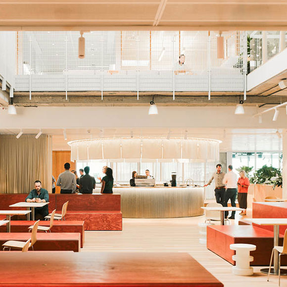 Workplace reception area with large central concierge, tables, chairs, sofas and plants set within warm tones.