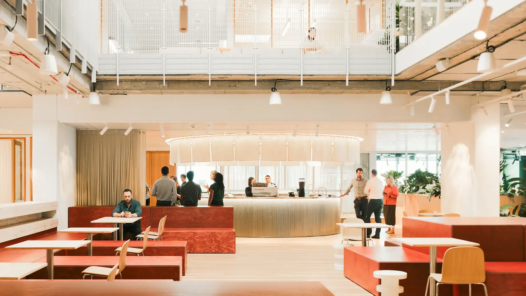 Workplace reception area with large central concierge, tables, chairs, sofas and plants set within warm tones.