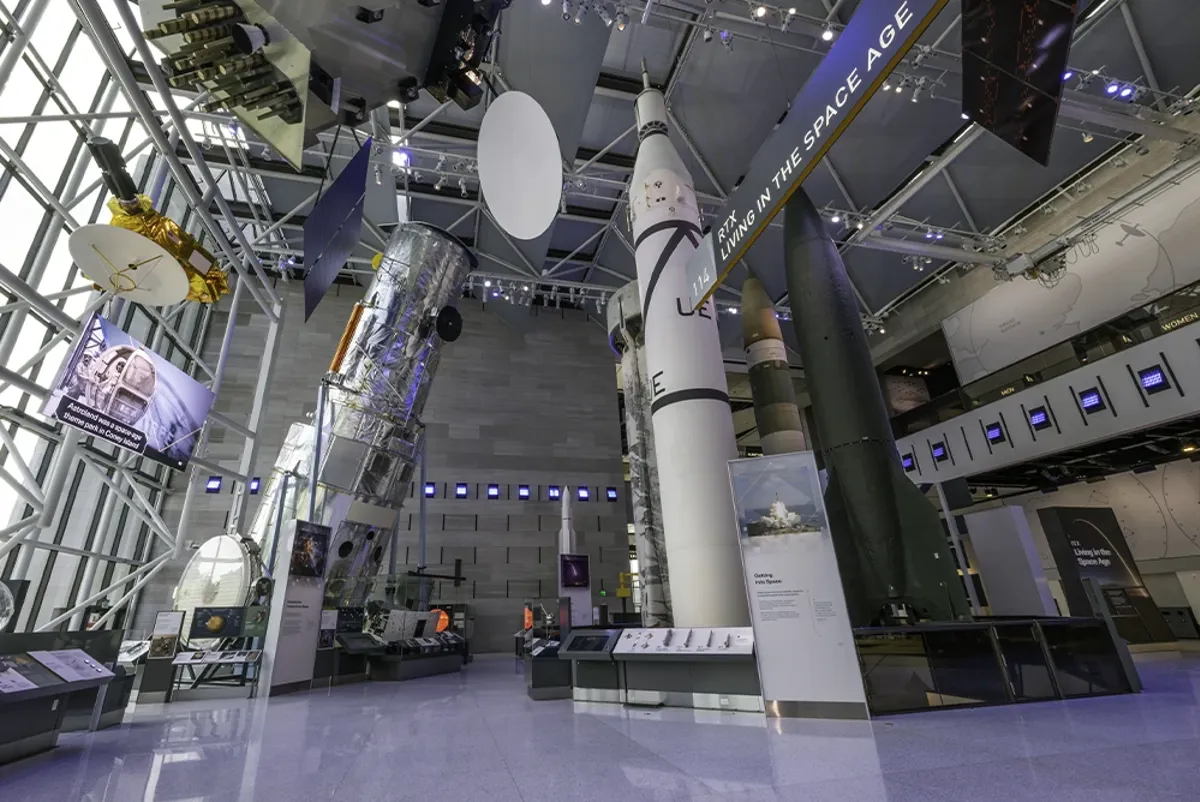 Space science artifacts including shuttles, rockets, and satellites hang from a system of exposed steel mounts in a room lit from light coming through a large window.