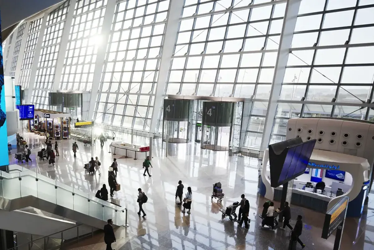 Passengers inside an airport terminal building with a large glass facade in the background